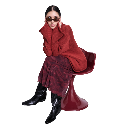 Woman in sunglasses, a red sweater, and a red patterned skirt sits on a modern red chair, resting her face in her hands, with black boots and hair pulled back.