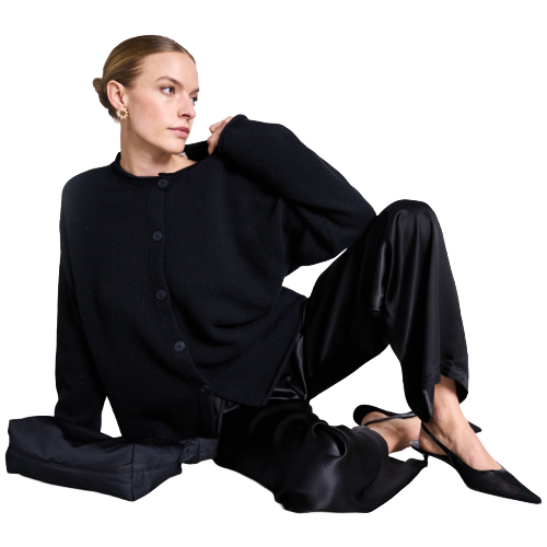 A woman in black clothing sits on the floor with one knee up and one arm resting on her raised knee, looking to the side. She wears pointed black heels and gold hoop earrings.