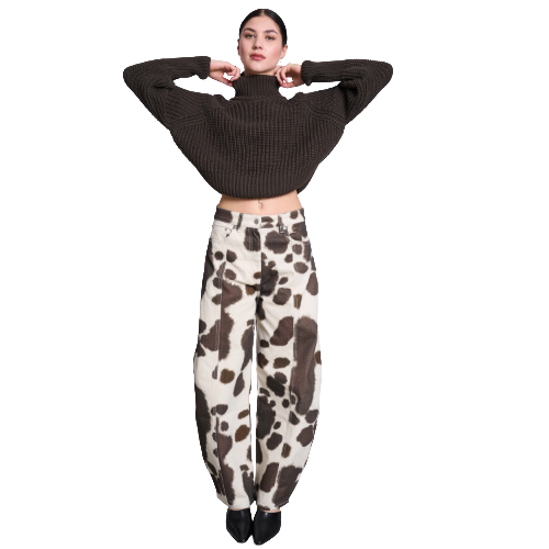 A woman stands with her arms raised, wearing a dark brown turtleneck sweater and loose, high-waisted cream-colored pants with a brown cow print pattern, paired with black shoes.