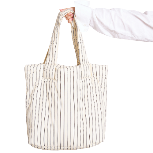 A hand in a white shirt holds a cream tote bag with vertical black stripes against a white background.