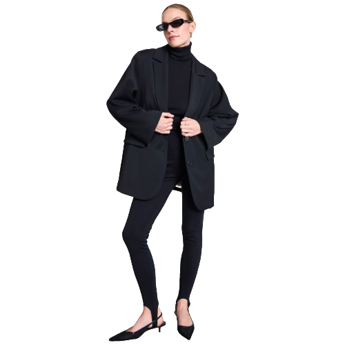 A woman poses in black sunglasses, a black oversized blazer, turtleneck, skinny pants, and pointed heels against a white background.