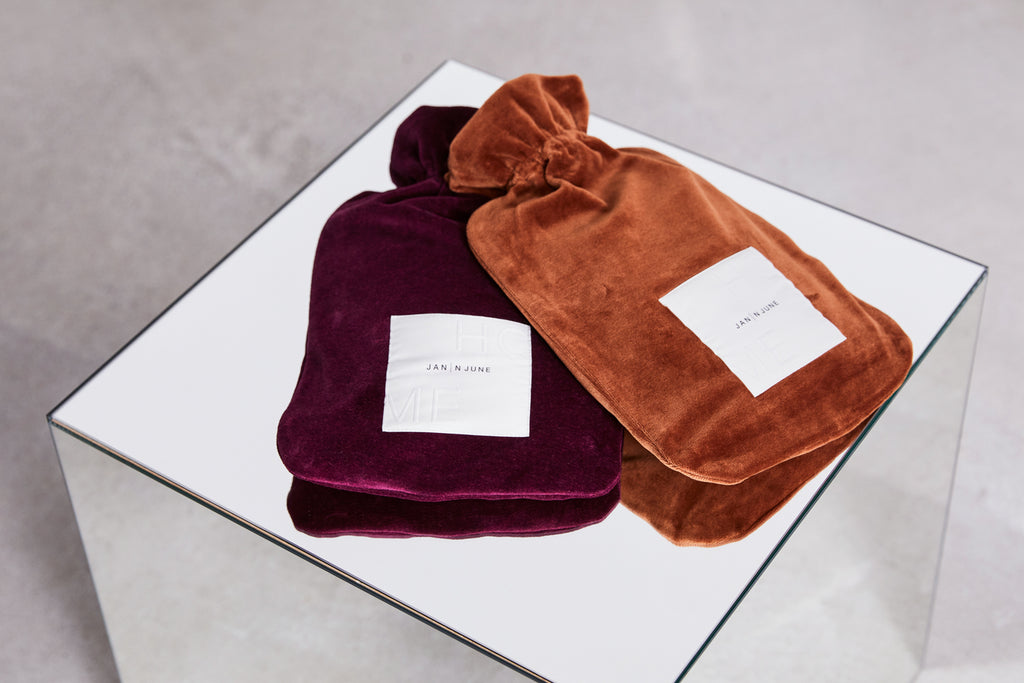 Two soft, velvet jan-n-june HOT WATER BOTTLE COVER NICKI BRICK in deep purple and rusty orange, each with a white logo label, sit on a mirrored table against a light gray background.