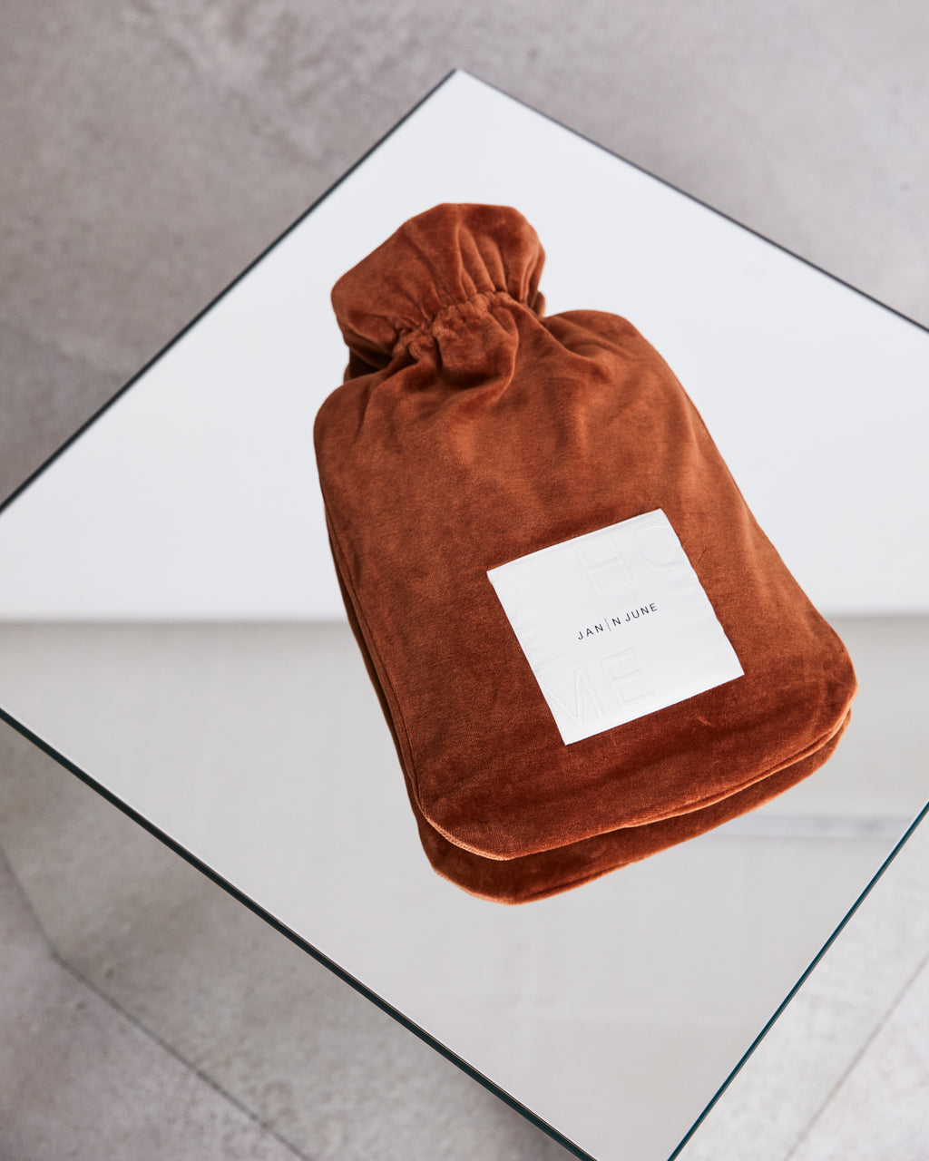 A rust-colored HOT WATER BOTTLE COVER NICKI BRICK by jan-n-june, with a white label, rests on a glass table over a grey floor—an example of our sustainable hot water bottle covers.
