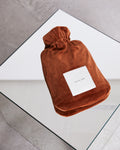 A rust-colored HOT WATER BOTTLE COVER NICKI BRICK by jan-n-june, with a white label, rests on a glass table over a grey floor—an example of our sustainable hot water bottle covers.