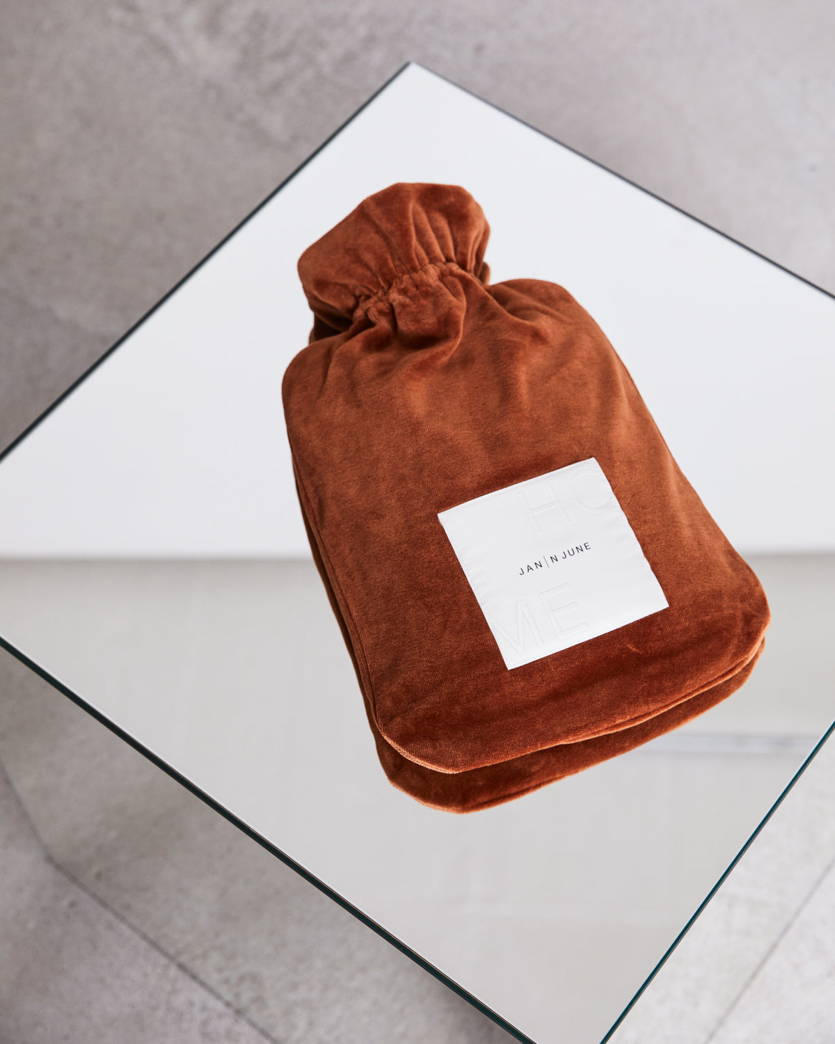 A rust-colored HOT WATER BOTTLE COVER NICKI BRICK by jan-n-june, with a white label, rests on a glass table over a grey floor—an example of our sustainable hot water bottle covers.