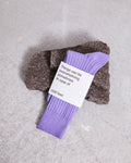A pair of comfy and soft SOCKS LILAC by jan-n-june, made from organic cotton, rests on a rough stone with a white label stating: “Things can be overwhelming sometimes. In case of cold feet.” Set against a light gray concrete background.