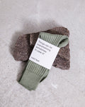 A pair of SOCKS SEA SPRAY by jan-n-june, made from green ribbed organic cotton, folded on a rough brown stone. The label reads, Things can be overwhelming sometimes. In case of cold feet. Set against a light textured background.