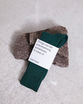 A pair of SOCKS BOTTLE GREEN by jan-n-june, made from organic cotton, rests on two rough stones. A white label says, Things can be overwhelming sometimes. In case of cold feet. Background: light and textured.
