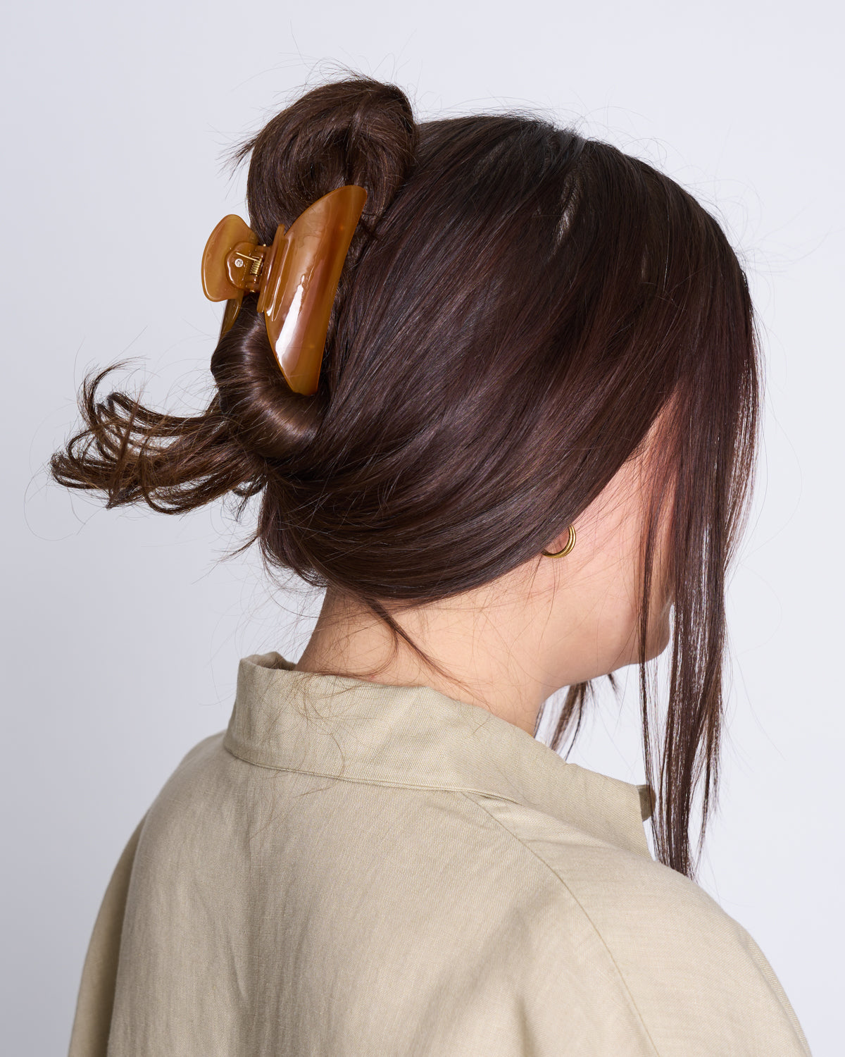A person with long brown hair in a twisted updo, secured by the jan-n-june MIDI HEIRLOOM CLAW COGNAC, wears a beige collared shirt and faces away from the camera against a plain light background.