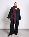 A woman stands against a white wall, wearing a black oversized suit, black shoes, glasses, and the jan-n-june SCARF ROSS CHERRY LACQUER in burgundy. She carries a large black tote bag and looks to the side with a neutral expression.