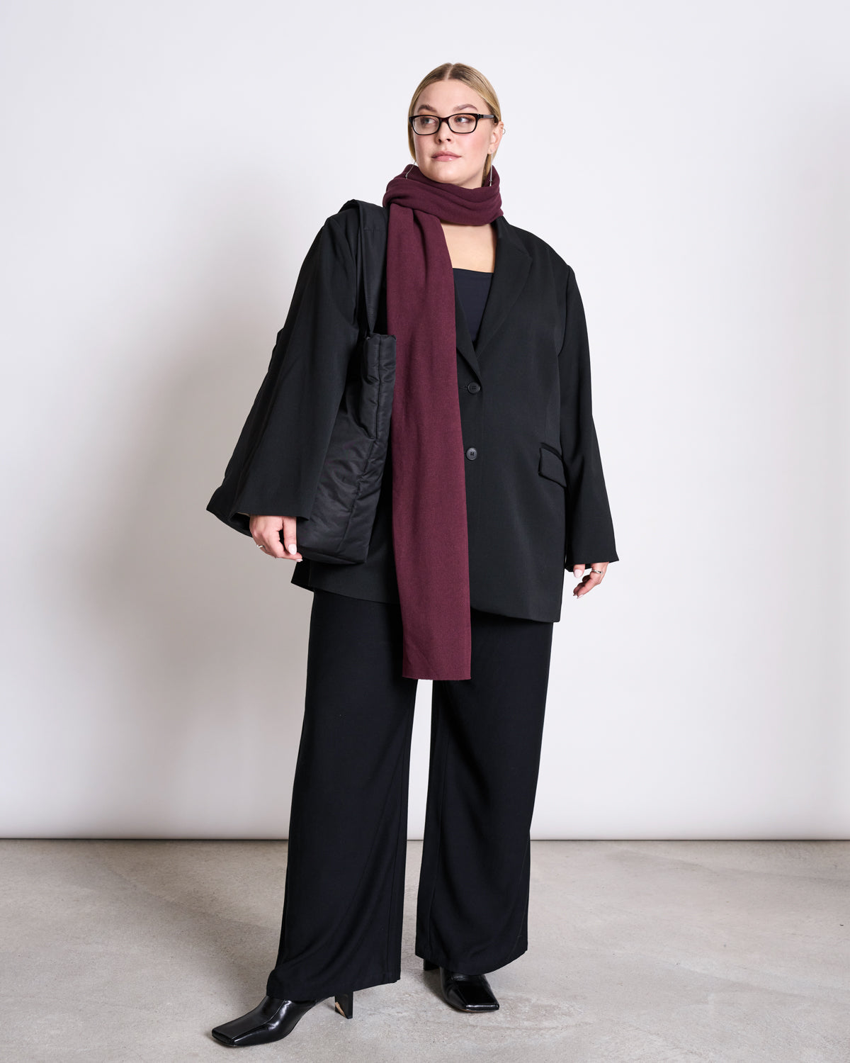 A woman stands against a white wall, wearing a black oversized suit, black shoes, glasses, and the jan-n-june SCARF ROSS CHERRY LACQUER in burgundy. She carries a large black tote bag and looks to the side with a neutral expression.