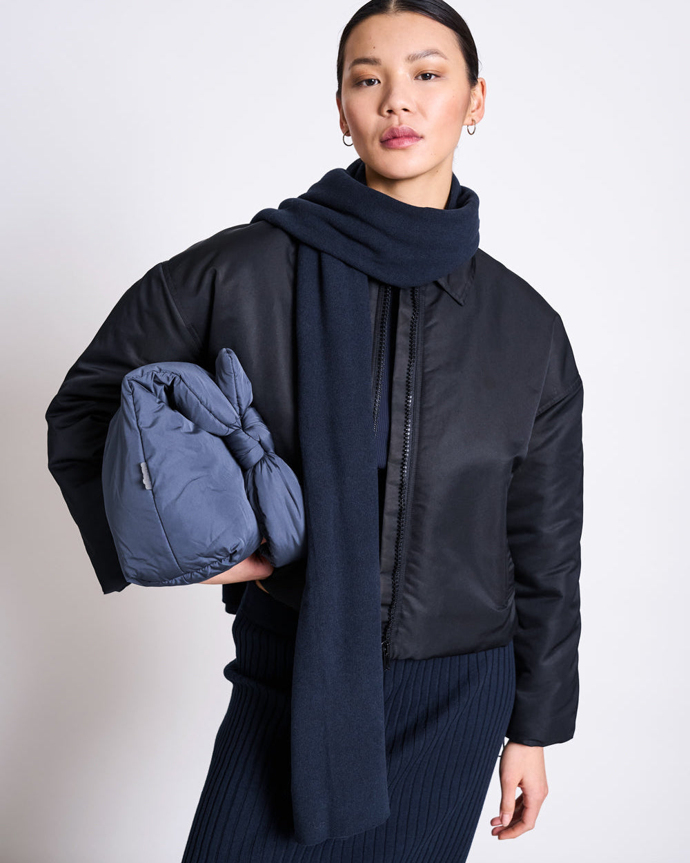 A person in a black jacket and ribbed skirt stands against a plain light background, holding a large blue knotted bag under one arm and wearing the jan-n-june SCARF ROSS NIGHTFALL navy wrap.