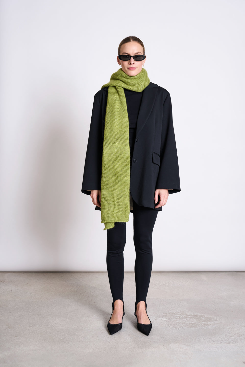 A person wearing black sunglasses, an oversized black blazer, leggings, and pointed shoes stands against a plain wall. Theyre wrapped in the jan-n-june WOOLY KNIT SCARF AVAK MOSSY PEAR, adding a cozy green accent to their look.