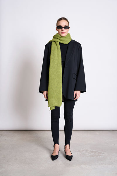 A person wearing black sunglasses, an oversized black blazer, leggings, and pointed shoes stands against a plain wall. Theyre wrapped in the jan-n-june WOOLY KNIT SCARF AVAK MOSSY PEAR, adding a cozy green accent to their look.