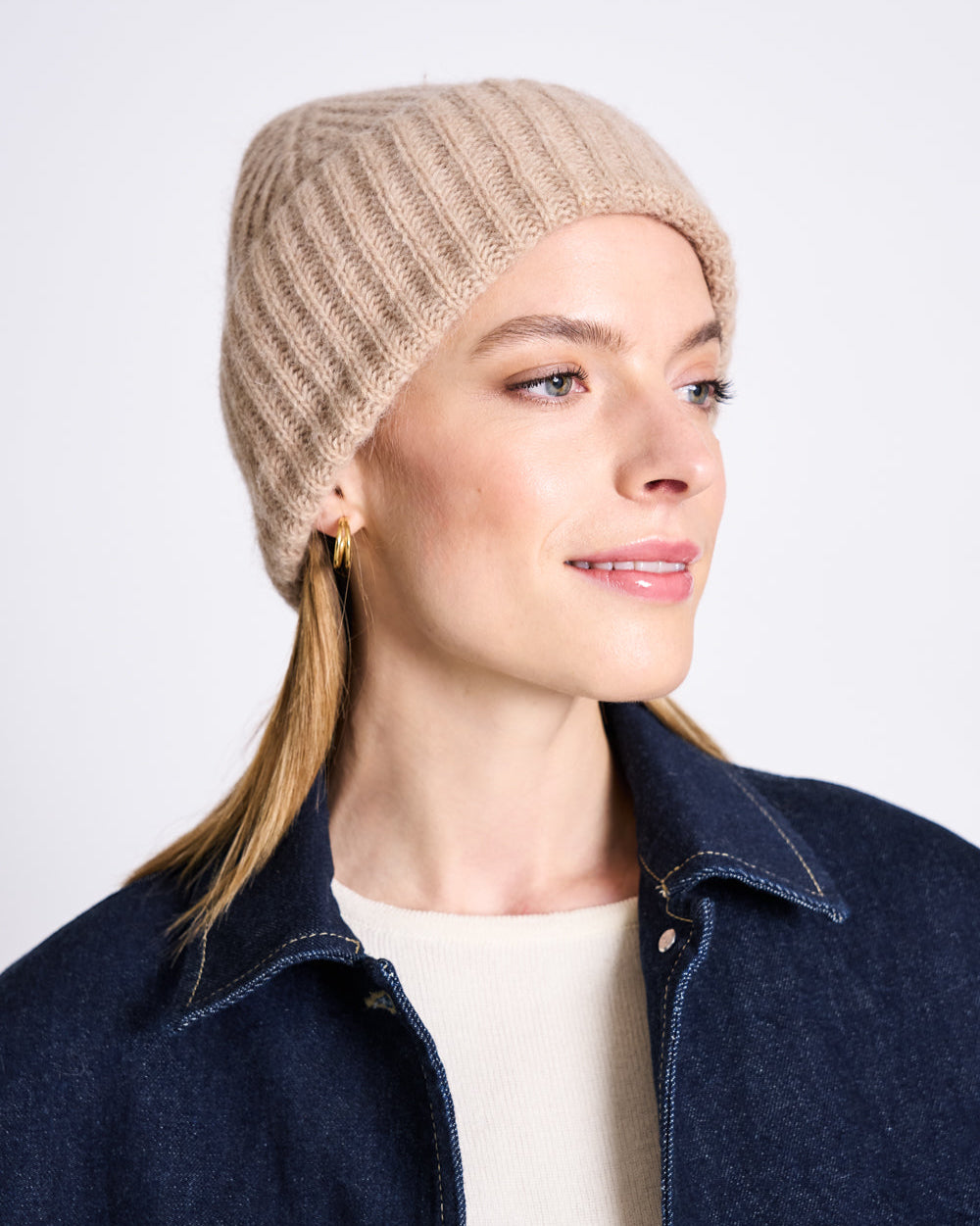 A woman wearing the jan-n-june WOOLY KNIT BEANIE BJON CAMEL with gold hoop earrings, a cream top, and a dark denim jacket smiles softly while looking slightly to the side against a light background.