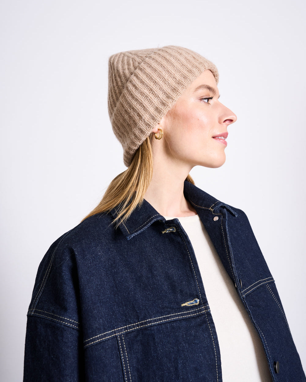 A woman in a beige WOOLY KNIT BEANIE BJON CAMEL by jan-n-june and gold hoop earrings looks to the side. She wears a dark denim jacket over a light top, with her blonde hair pulled back. The beanie is offered in various colors. Plain white background.