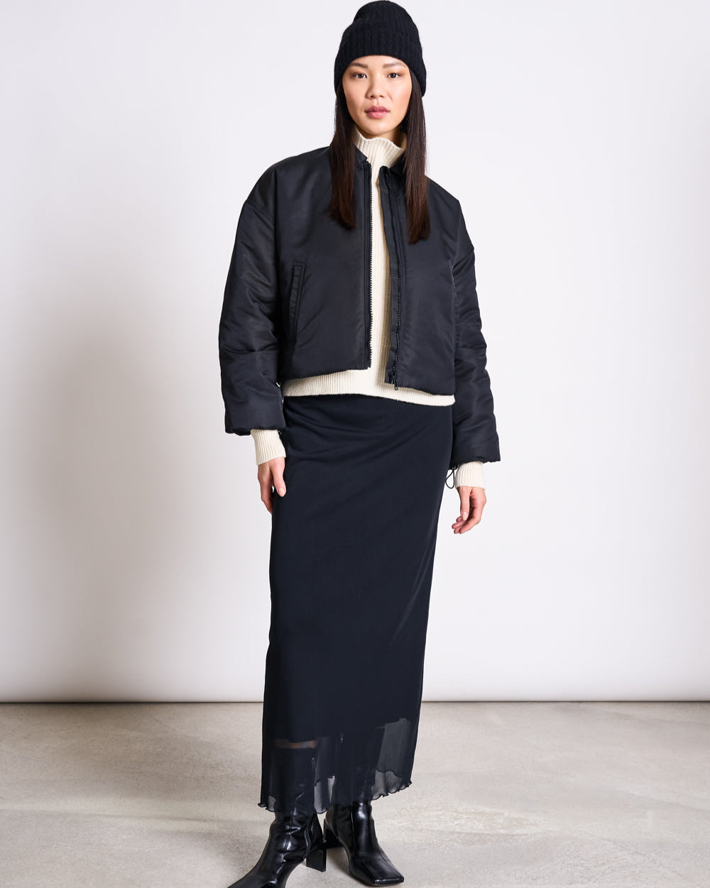 A woman stands against a plain background, wearing the jan-n-june WOOLY KNIT BEANIE BJON BLACK, a black bomber jacket, cream turtleneck sweater, long black skirt, and black ankle boots. She looks at the camera neutrally. The beanie is available in various colors.