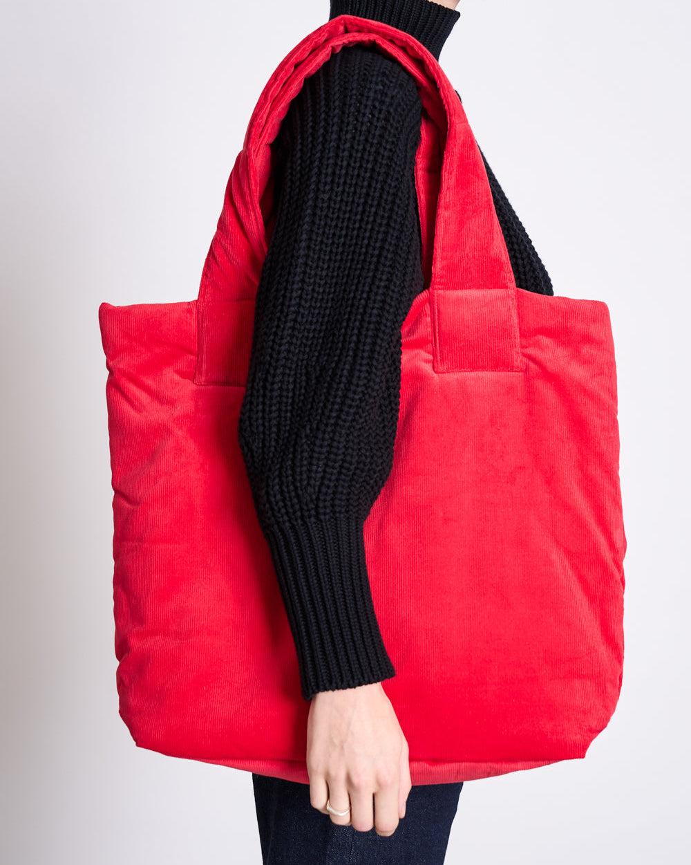 A person wears a black knit sweater and carries the jan-n-june PADDED SHOPPER SMOL RED, a bright red deadstock shopper, over their shoulder. The cropped image shows the upper body and bag against a plain background.