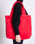 A person wears a black knit sweater and carries the jan-n-june PADDED SHOPPER SMOL RED, a bright red deadstock shopper, over their shoulder. The cropped image shows the upper body and bag against a plain background.