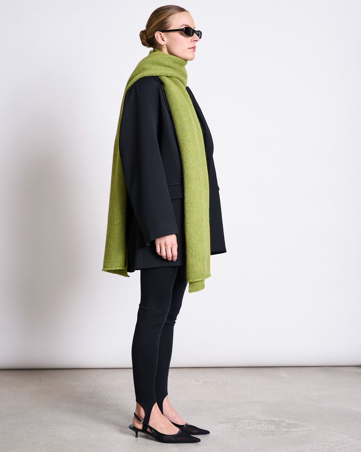A woman in profile wears a black blazer, leggings, slingback shoes, sunglasses, and the jan-n-june WOOLY KNIT SCARF AVAK MOSSY PEAR draped around her neck, set against a plain white background.