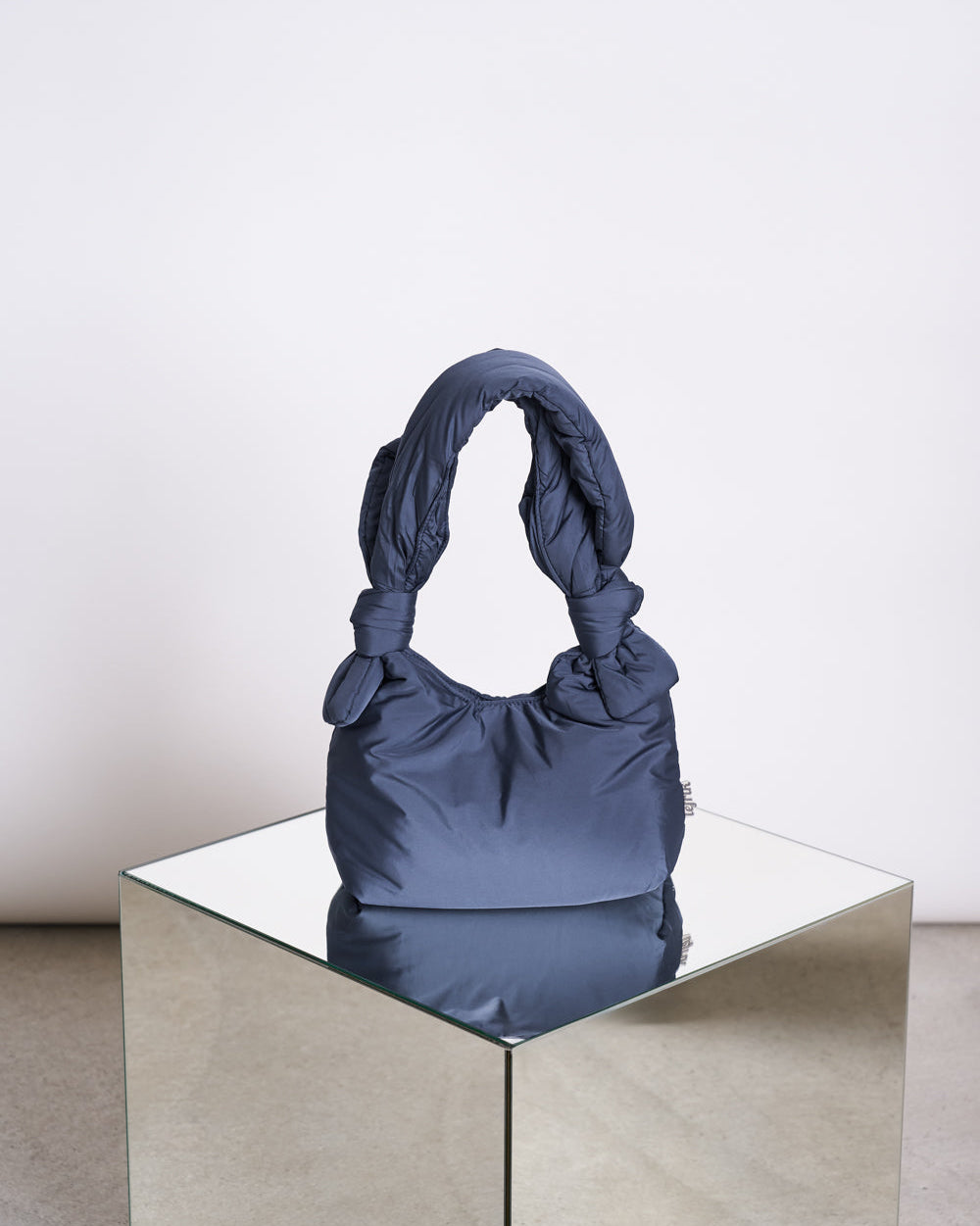 The jan-n-june BAG BIWA PUFFY MINI MAIBON, a dark blue mini shoulder bag with a knotted handle in quilted RPET fabric, is displayed on a reflective glass cube against a plain white background.