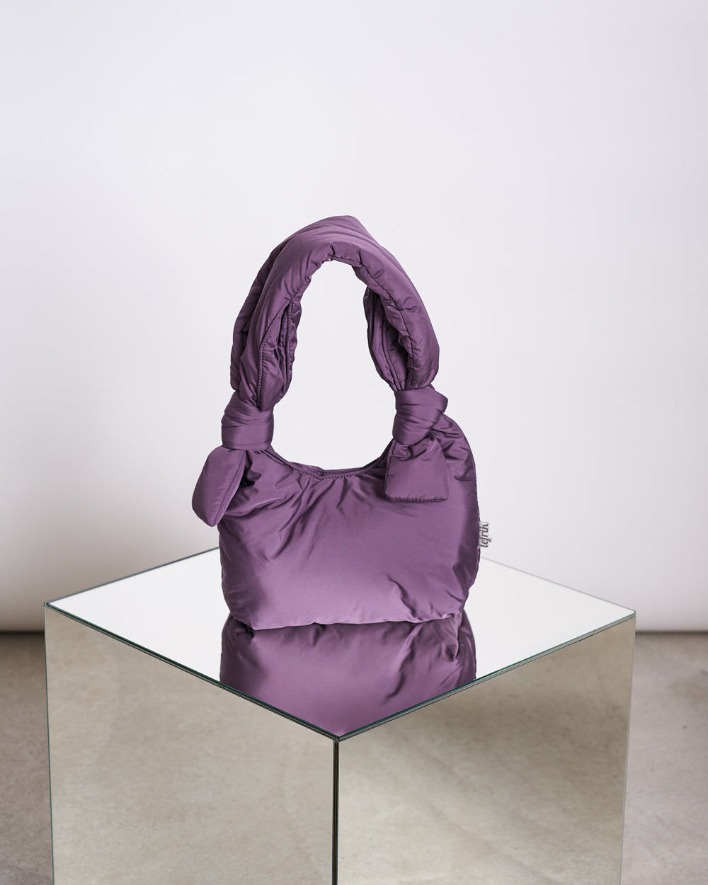 The jan-n-june BAG BIWA PUFFY MINI PLUM—a soft, quilted RPET bag in plum with knotted handles—rests on a mirrored cube against a plain, light background.