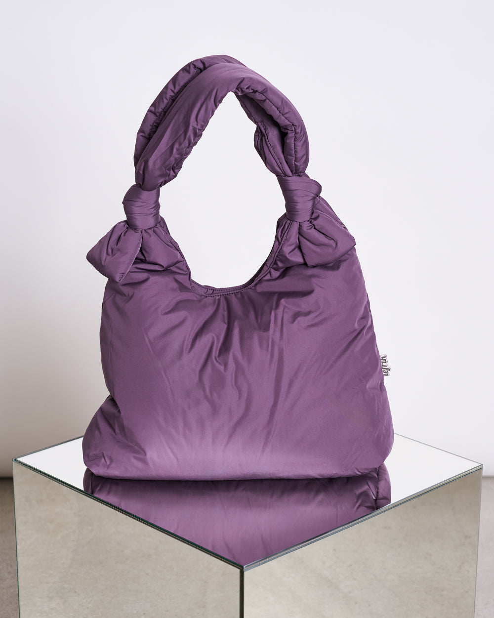 The BAG BIWA PUFFY REGULAR PLUM by jan-n-june is a purple, quilted vegan bag with knotted handles, crafted from recycled plastic. It sits on a mirrored surface against a light background, blending style and sustainability.