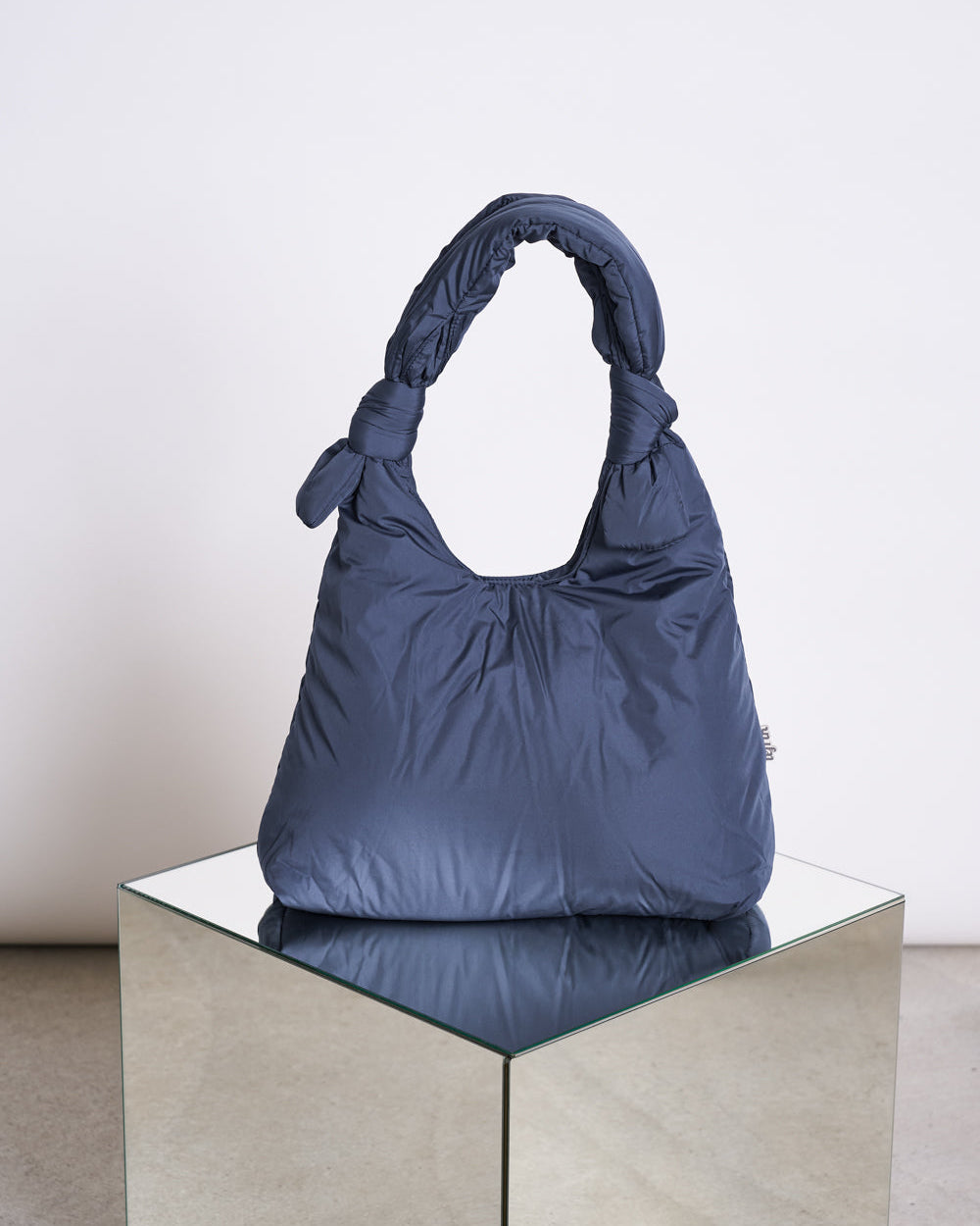 The BAG BIWA PUFFY REGULAR MAIBON by jan-n-june, a dark blue quilted bag with knotted handles, is displayed on a reflective square mirrored pedestal against a plain light background.