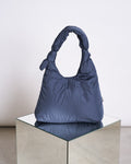 The BAG BIWA PUFFY REGULAR MAIBON by jan-n-june, a dark blue quilted bag with knotted handles, is displayed on a reflective square mirrored pedestal against a plain light background.