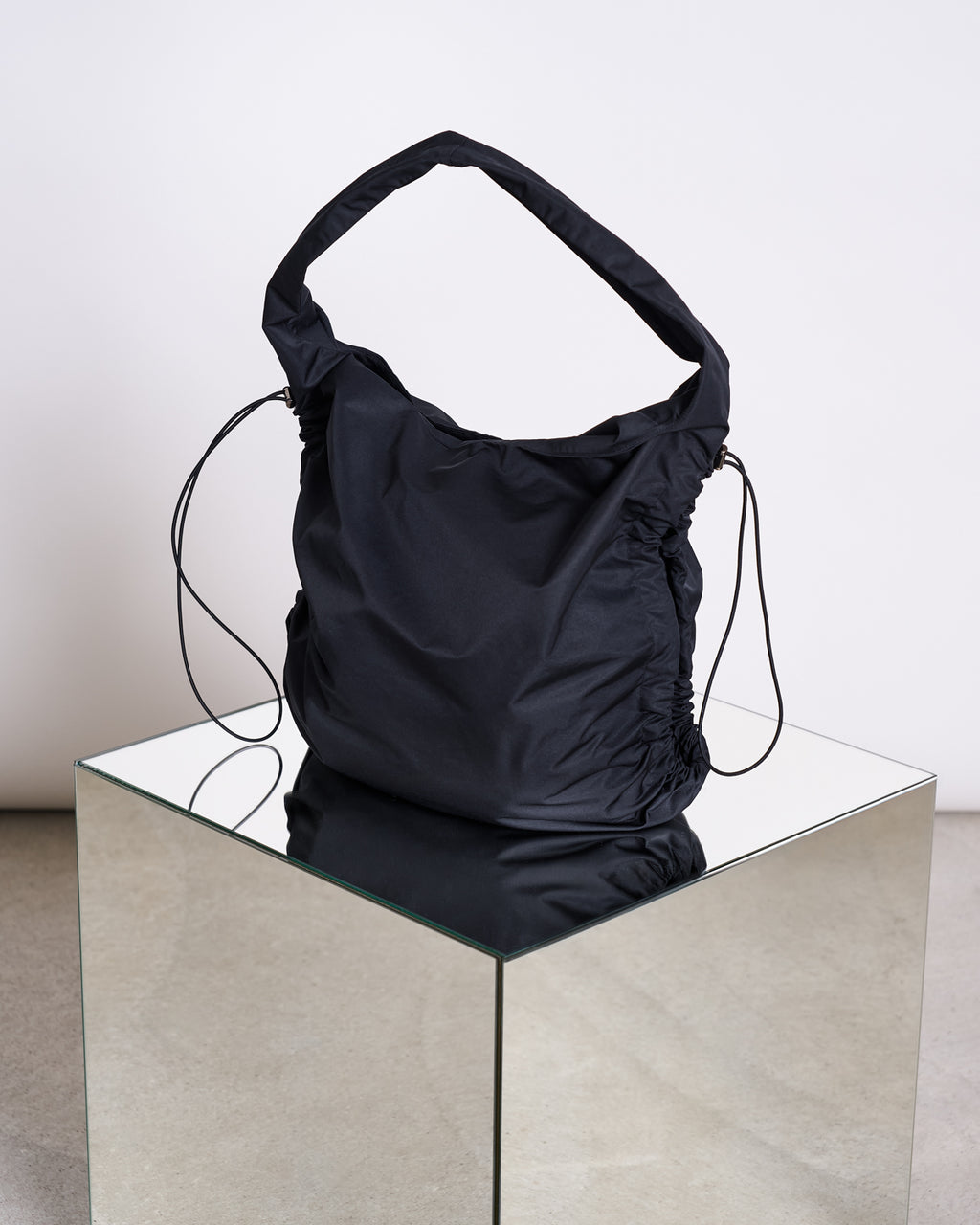 The jan-n-june DRAWSTRING SHOPPER LYNE BLACK, made from recycled polyester with ruched sides and slim drawstring details, is displayed on a reflective glass cube against a neutral background.