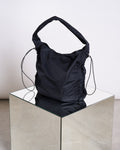 The jan-n-june DRAWSTRING SHOPPER LYNE BLACK, made from recycled polyester with ruched sides and slim drawstring details, is displayed on a reflective glass cube against a neutral background.