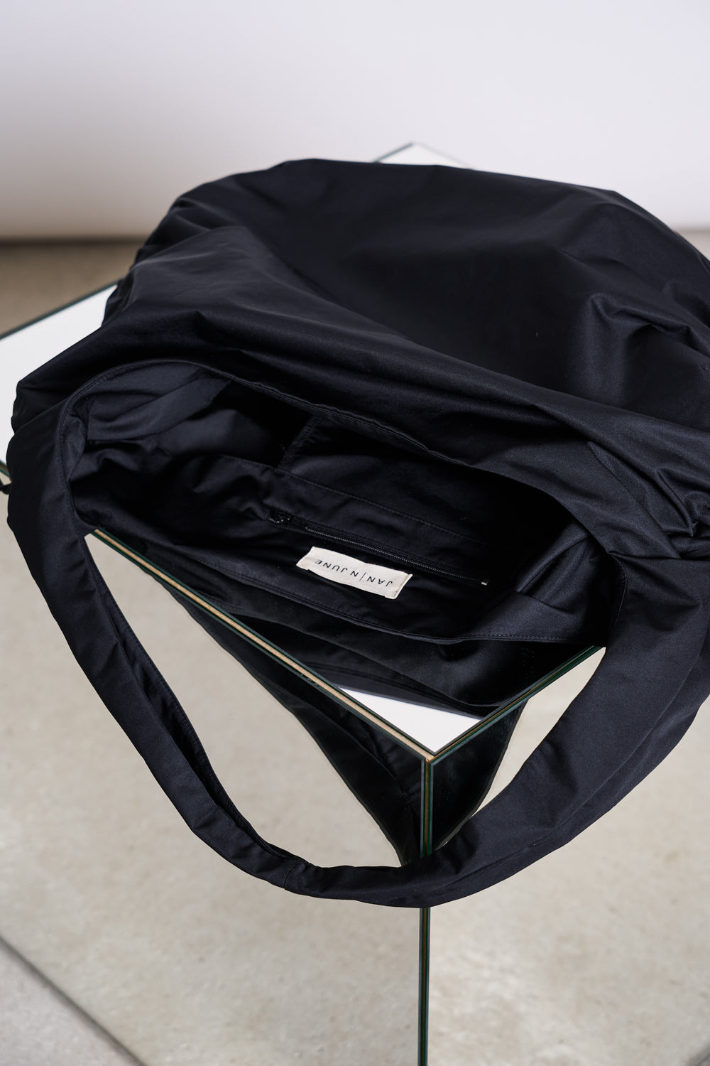A jan-n-june DRAWSTRING SHOPPER LYNE BLACK tote, crafted from recycled polyester with a white “SANNIN” label, sits open on a glass table showing its inner pocket and handle, set against a light grey floor and white wall.