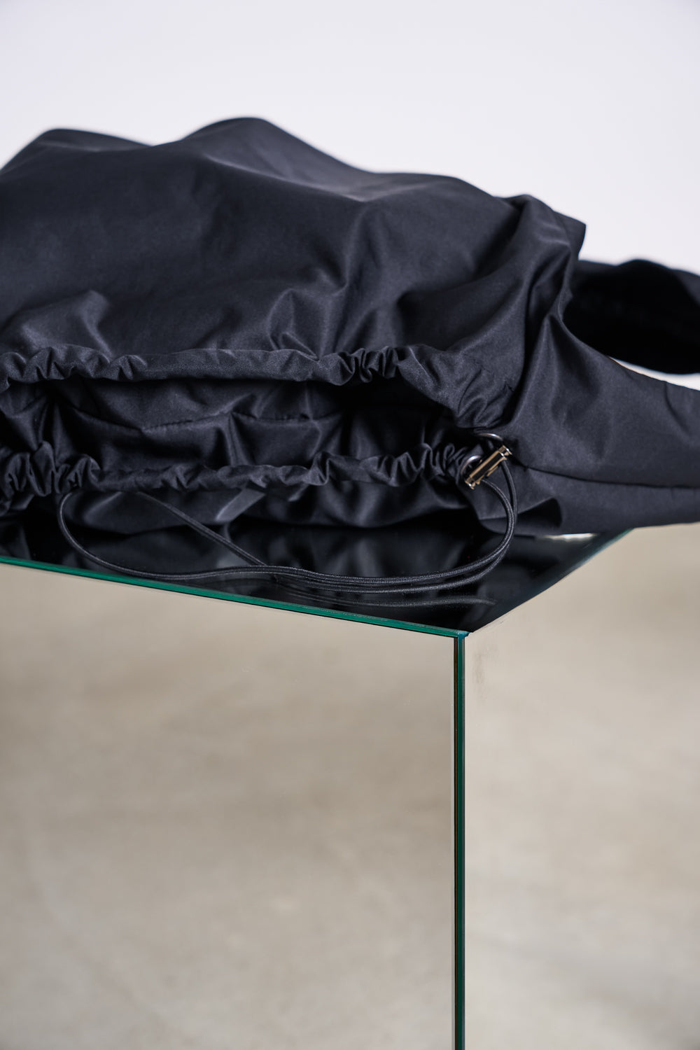 The DRAWSTRING SHOPPER LYNE BLACK by jan-n-june, made from recycled polyester with gathered fabric details, sits on the edge of a glass table against a neutral background.