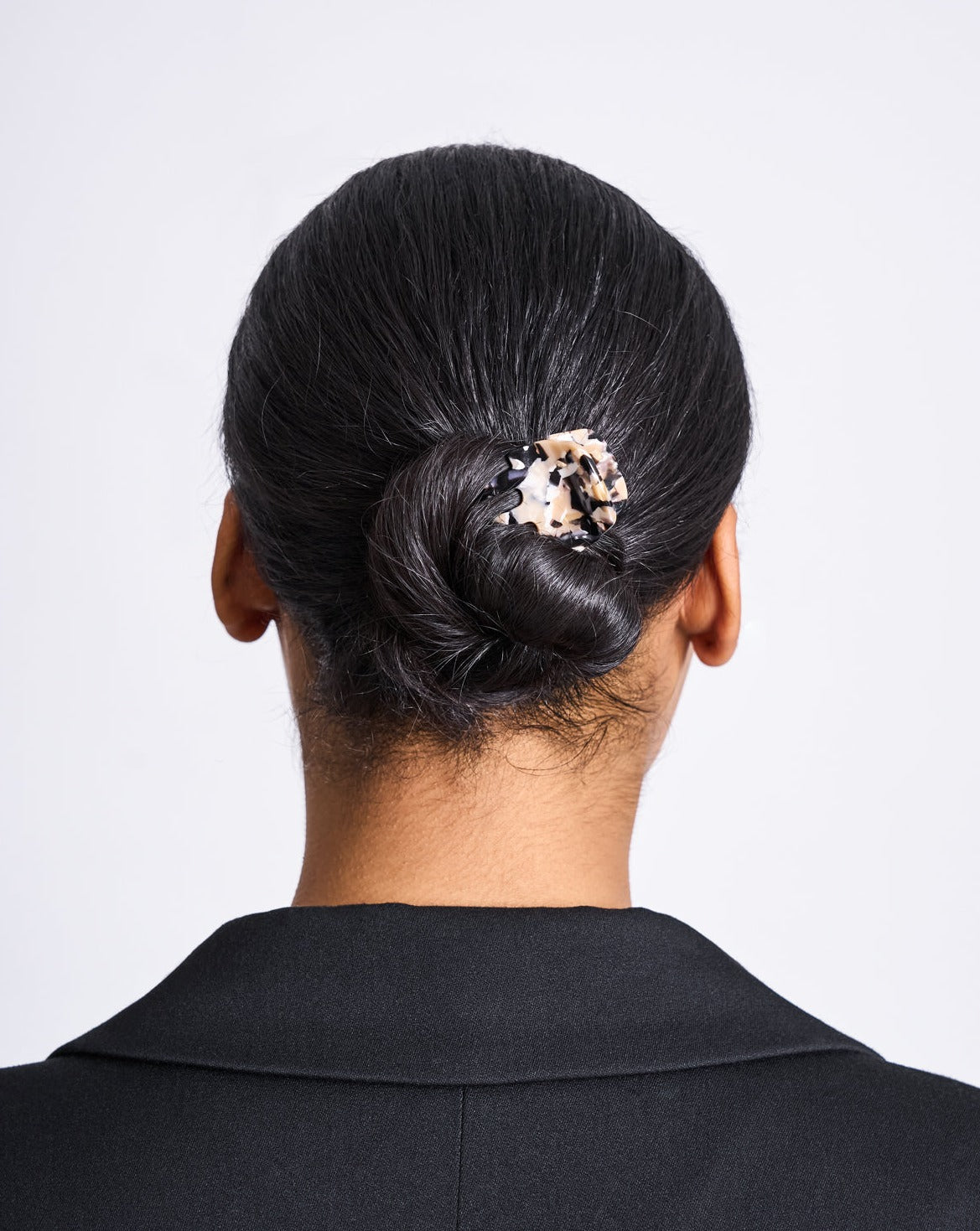A person with dark hair in a low twisted bun secured by the jan-n-june MINI CLAW ABALONE handcrafted acetate hair clip, wearing a black collared top, shown from the back against a plain light background.