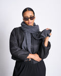A person in dark sunglasses and a black jacket stands confidently against a plain background, holding the end of the jan-n-june SCARF ROSS DARK GREY, a cozy scarf made from recycled cotton.