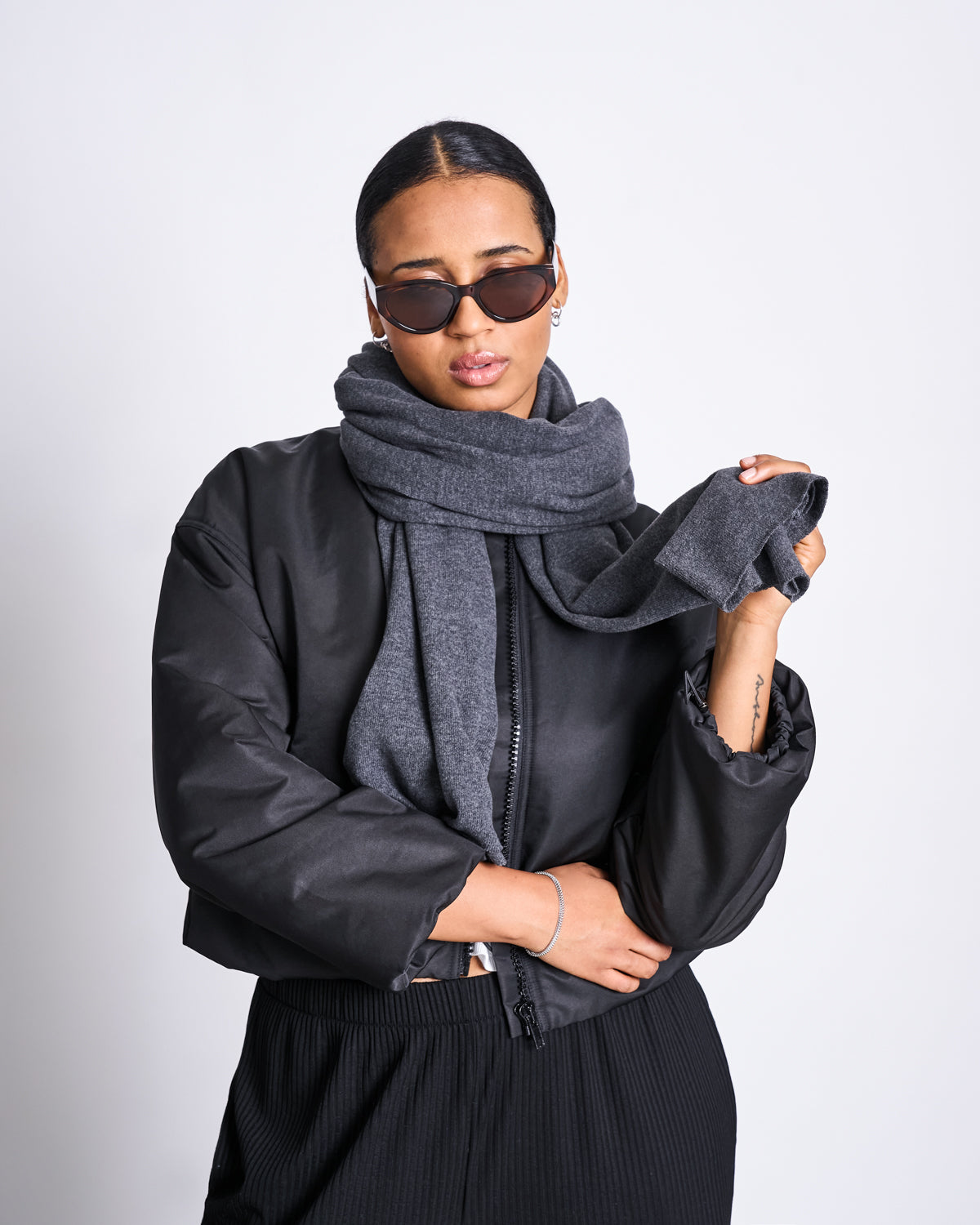 A person in dark sunglasses and a black jacket stands confidently against a plain background, holding the end of the jan-n-june SCARF ROSS DARK GREY, a cozy scarf made from recycled cotton.