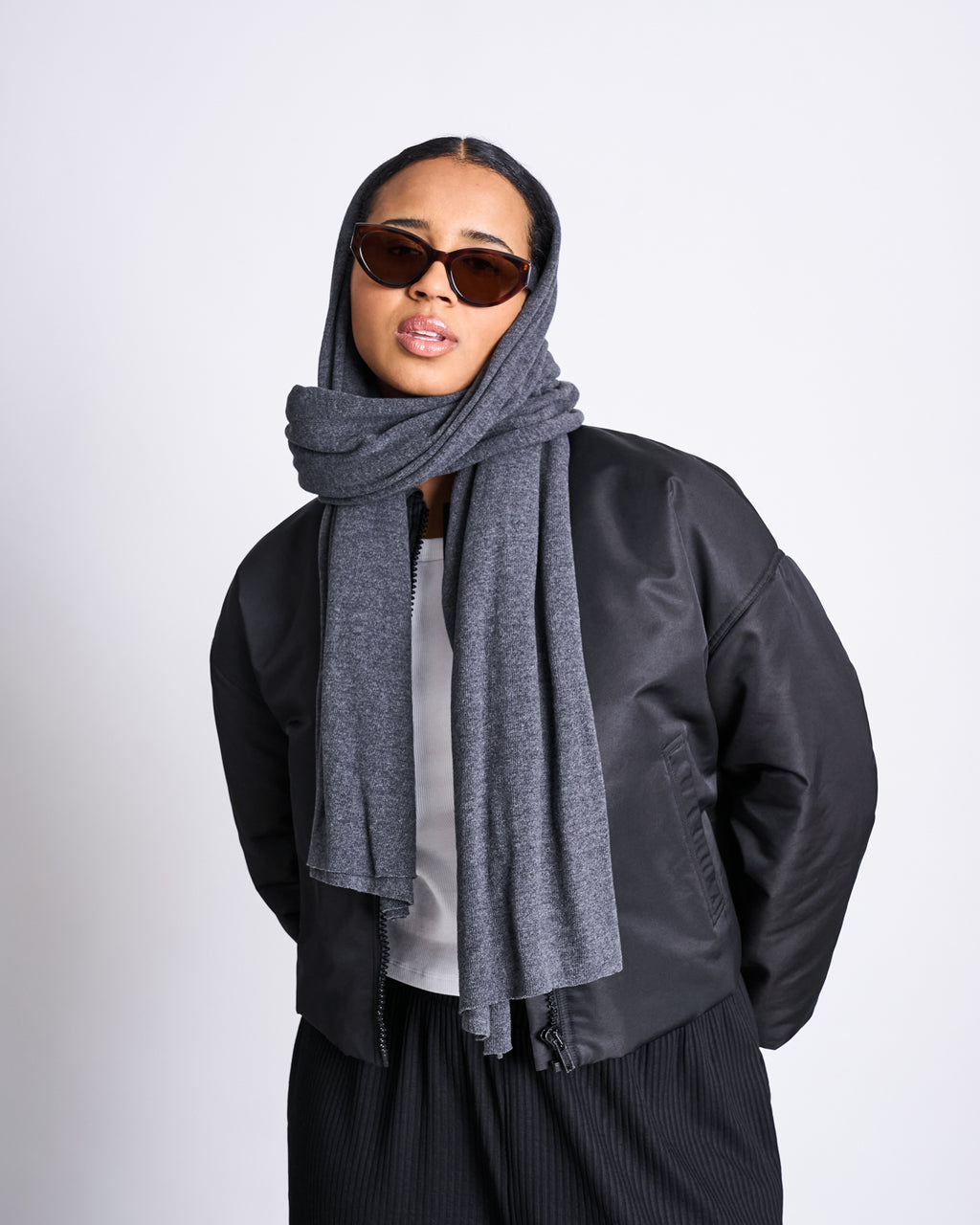 A person models the jan-n-june SCARF ROSS DARK GREY wrapped around their head, paired with dark sunglasses, a black jacket, white top, and black pants against a plain light background.