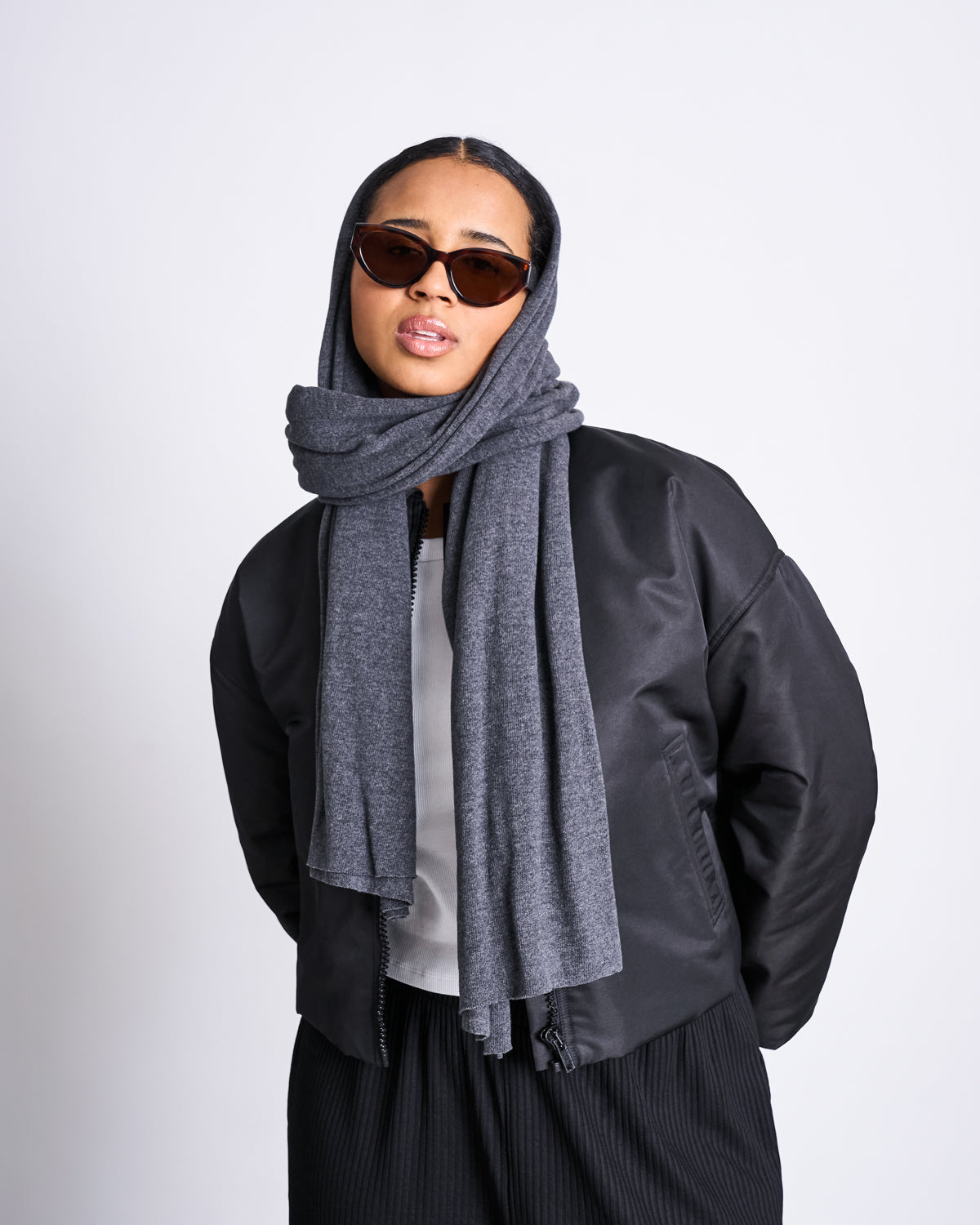 A person models the jan-n-june SCARF ROSS DARK GREY wrapped around their head, paired with dark sunglasses, a black jacket, white top, and black pants against a plain light background.
