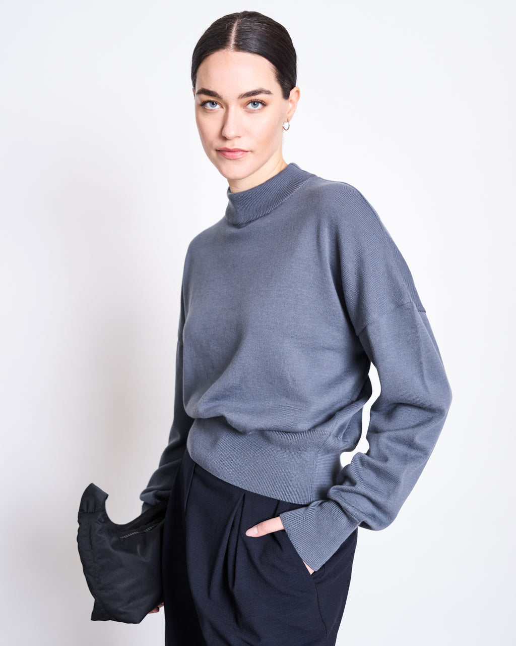 A woman with dark hair in a low bun wears the jan-n-june JUMPER YIN TURBULENCE GOTS organic cotton knit and dark pants, standing against a white background holding a black accessory in her left hand.