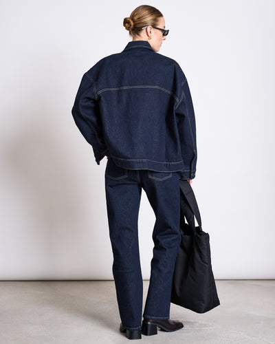 A person with blonde hair in a bun wears the jan-n-june RELAXED DENIM JACKET VILME DARK BLUE DENIM GOTS, dark blue jeans, black boots, and sunglasses while holding a black tote bag against a plain background.