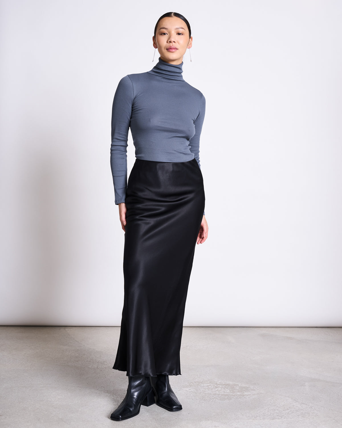A woman stands indoors in the jan-n-june MINIRIB TURTLENECK CHUVA TURBULENCE GOTS, paired with a long black satin skirt and ankle boots, her hair pulled back as she poses against a plain white wall.