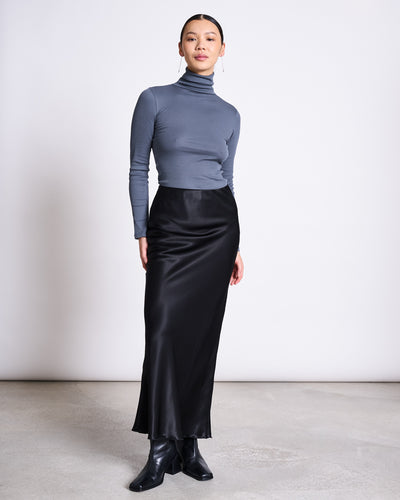 A woman poses confidently in a minimal studio, wearing the jan-n-june MAXI SKIRT SERIN BLACK with a fitted gray turtleneck and black ankle boots. Her hair is slicked back as she stands against a plain white background.