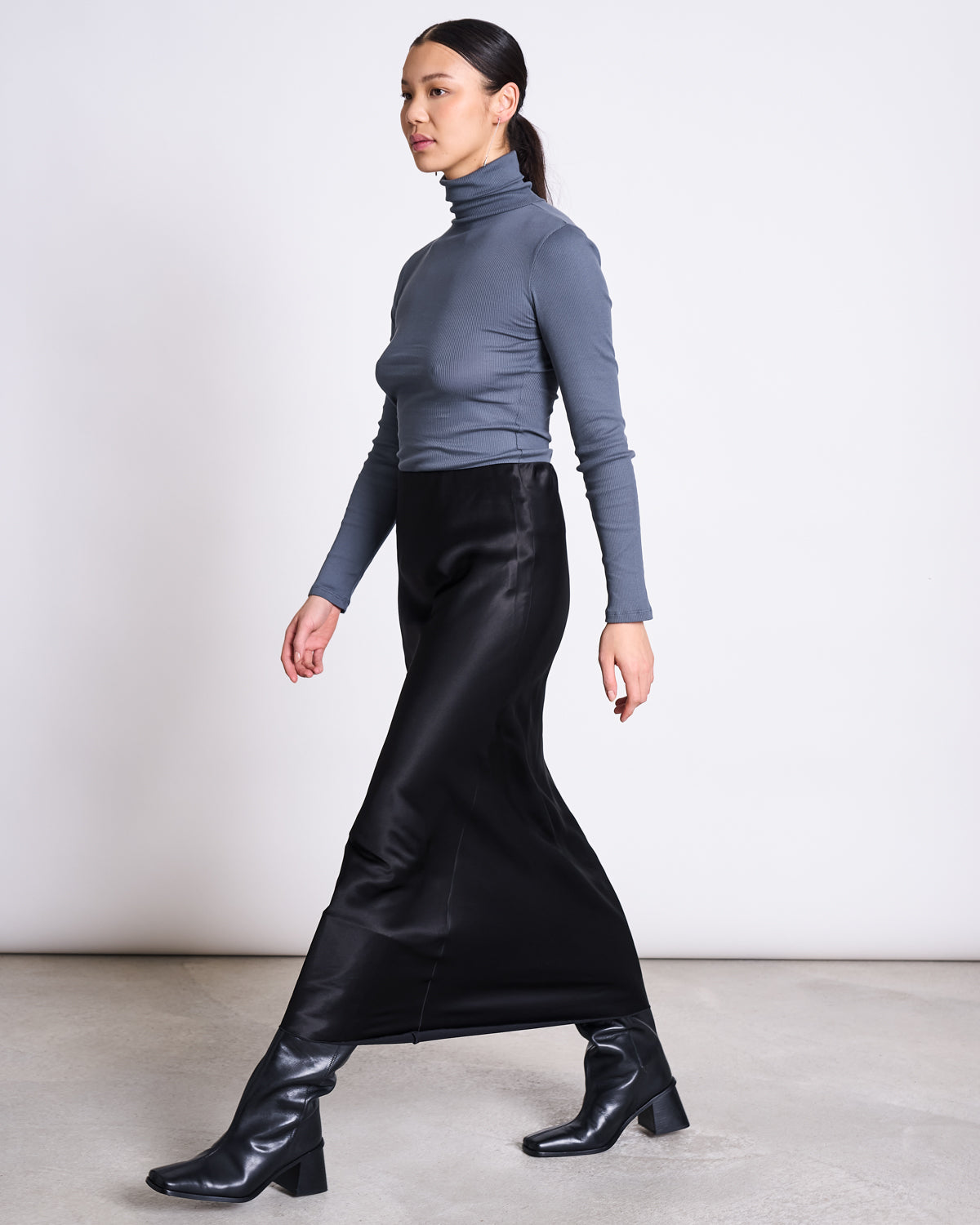 A woman with long dark hair in a ponytail walks indoors, wearing the jan-n-june MINIRIB TURTLENECK CHUVA TURBULENCE GOTS, a slim fit gray turtleneck, against a plain white background.