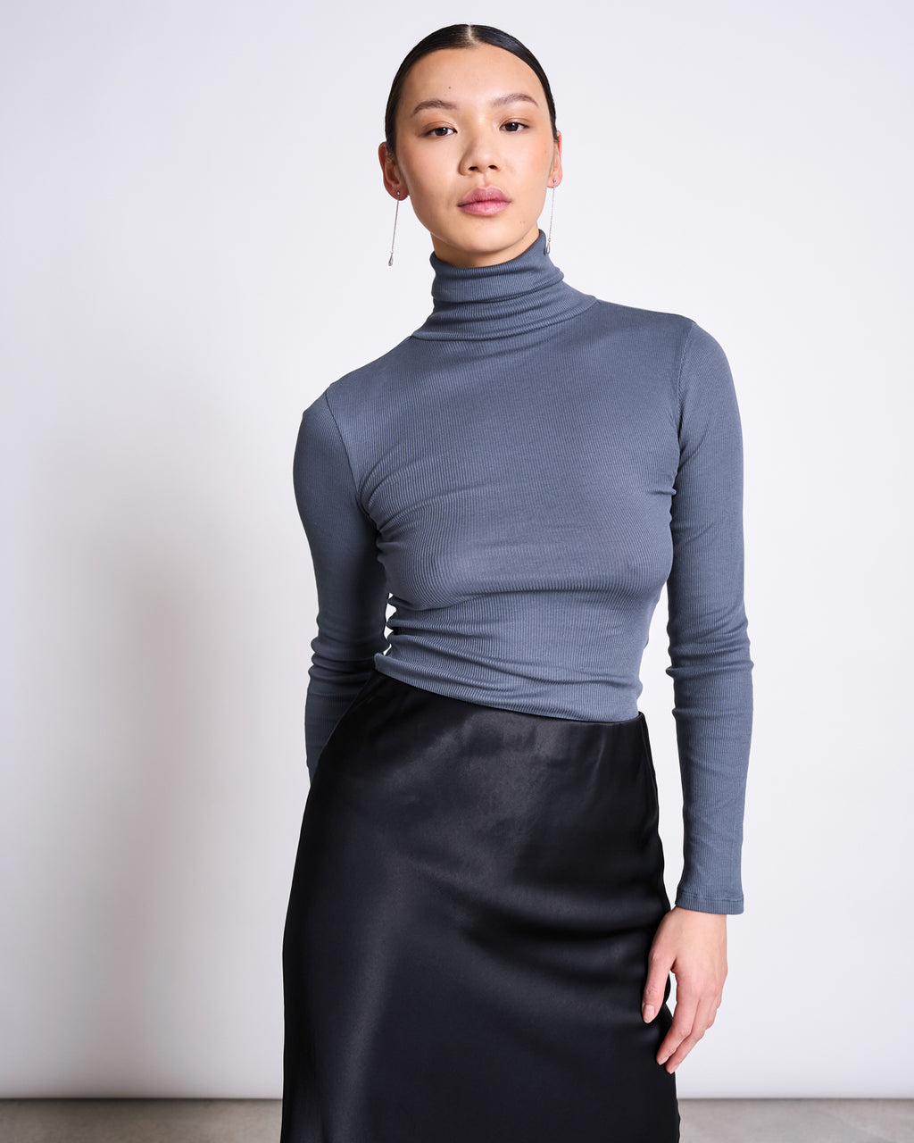 A woman stands against a plain light background, wearing the jan-n-june MINIRIB TURTLENECK CHUVA TURBULENCE GOTS in gray with a black satin skirt; her hair is slicked back and one hand rests by her side.