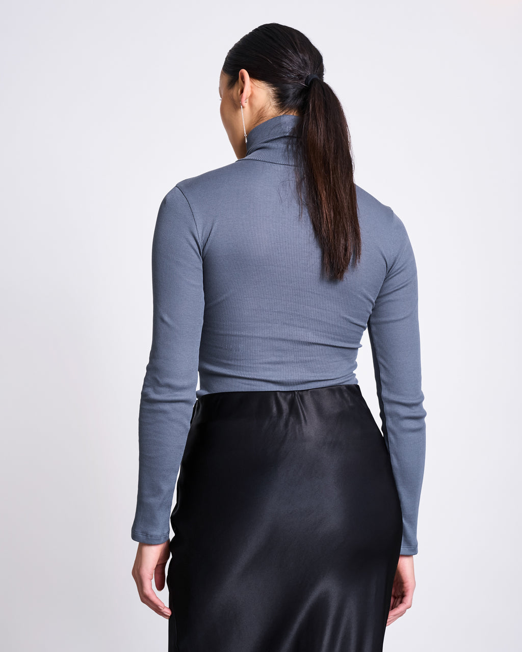 A woman with long dark hair in a low ponytail is seen from the back, wearing the jan-n-june MINIRIB TURTLENECK CHUVA TURBULENCE GOTS slim-fit gray top and a black satin skirt against a plain white background.