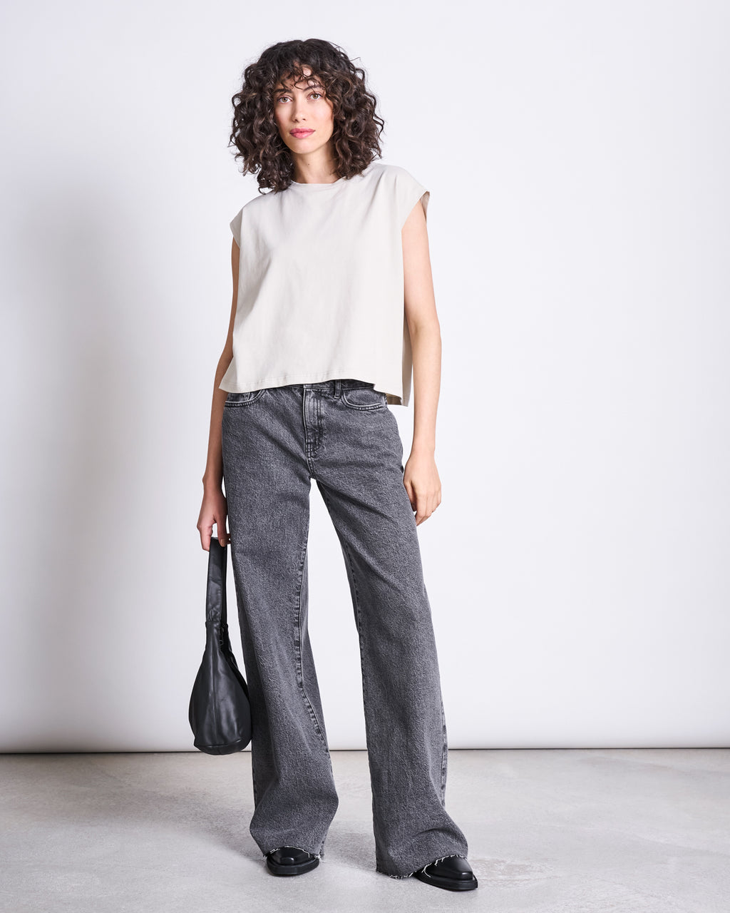 A woman with curly hair wears the jan-n-june WIDE LEG JEANS SUNNY LIGHT GREY, a sleeveless beige top, black shoes, and holds a black handbag at her side, standing against a plain white background.