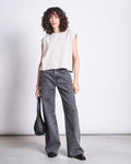 A woman with curly hair wears the jan-n-june WIDE LEG JEANS SUNNY LIGHT GREY, a sleeveless beige top, black shoes, and holds a black handbag at her side, standing against a plain white background.