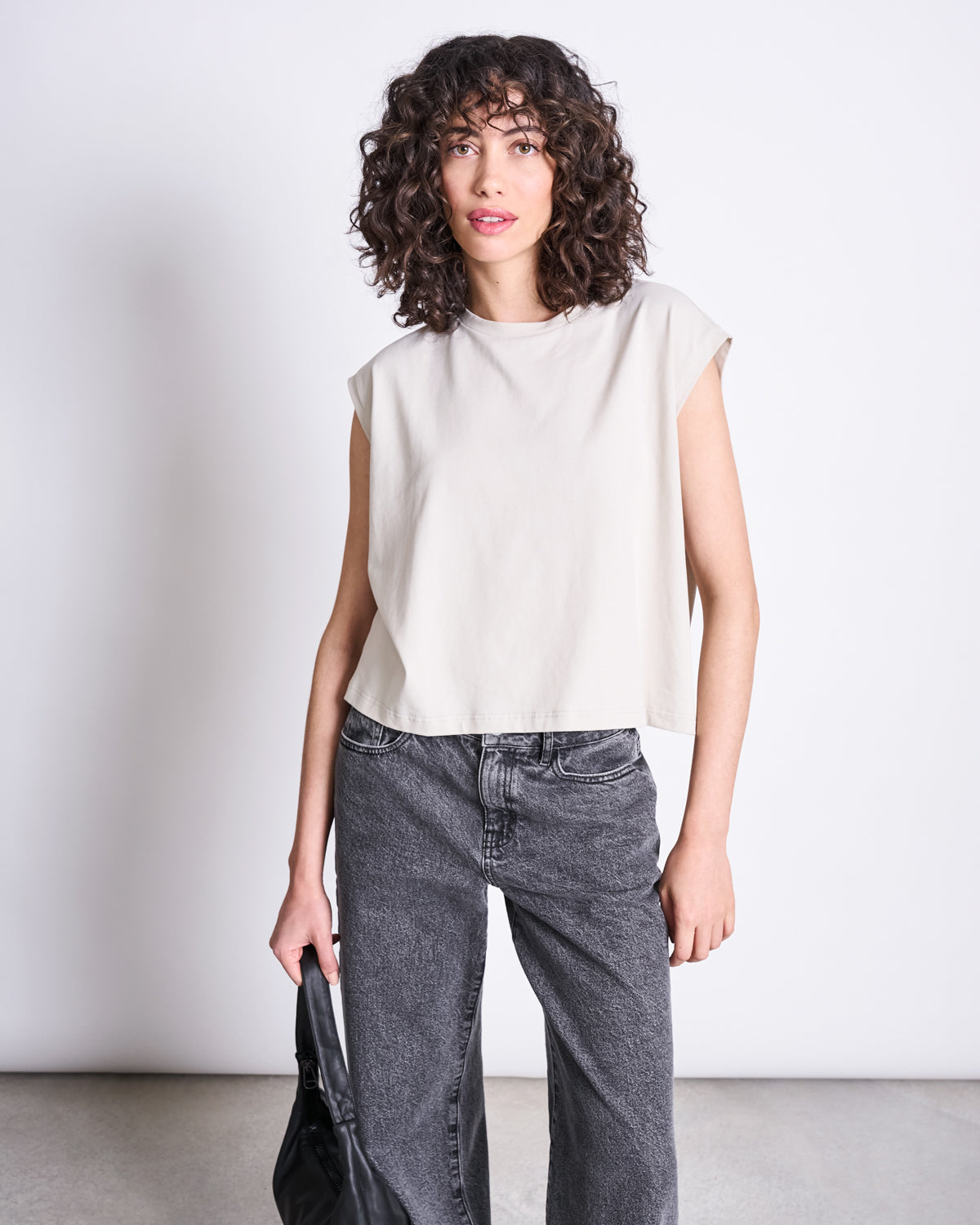 A woman with curly hair wears jan-n-junes WIDE LEG JEANS SUNNY LIGHT GREY and a sleeveless off-white top, standing against a plain background and holding a black bag at her side.