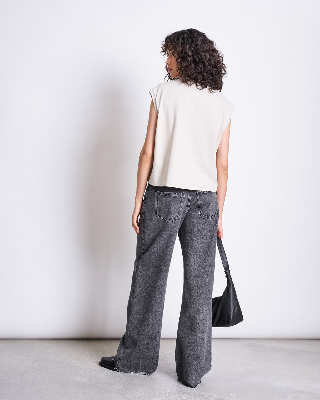 A person with curly hair faces away, wearing a sleeveless beige top, jan-n-junes WIDE LEG JEANS SUNNY LIGHT GREY known for high comfort, and black shoes. They hold a black handbag by their side against a plain white background.