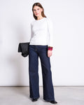 A woman with straight brown hair wears the jan-n-june RIB LONGSLEEVE RAVA WHITE top, dark blue wide-leg jeans, black shoes, and holds a black jacket. She stands against a white wall and accessorizes with colorful bangles.